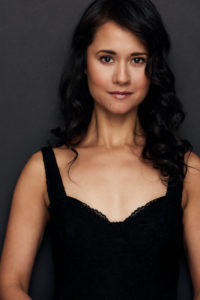 Actor Ali Ewoldt, self-identified as a female Filipino-American looks at the camera with a content face, wearing a plain black tank-top styled blouse and her hair naturally curly down to her shoulders.